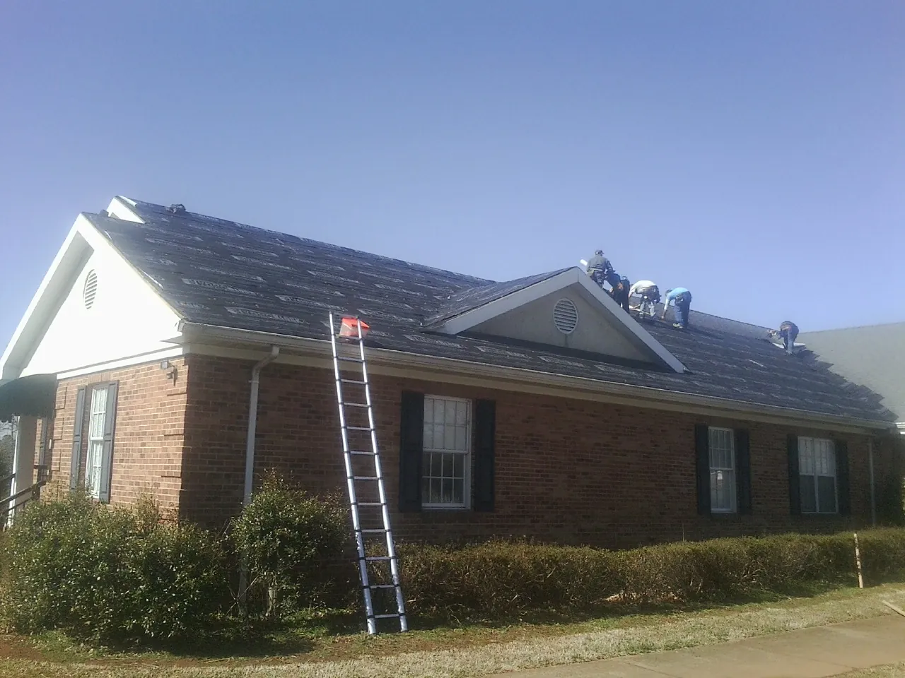 Crew tearing shingles off a brick ranch roof with a ladder set — Emergency Roof Repair work by our crew in Corolla