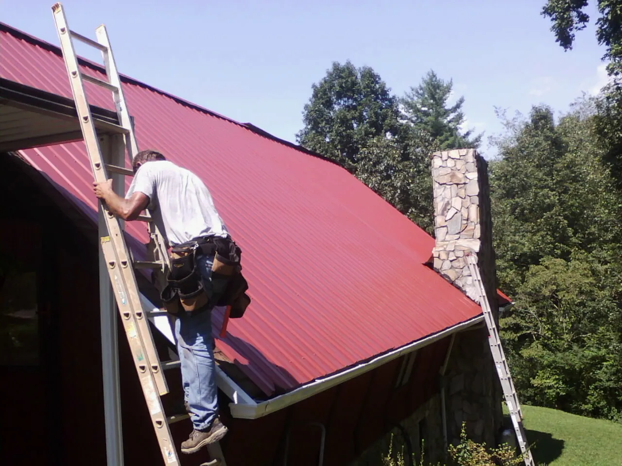 Roofer on a ladder beside a red metal roof and stone chimney — Soffit & Fascia Repair by Roofing Brothers in Corolla